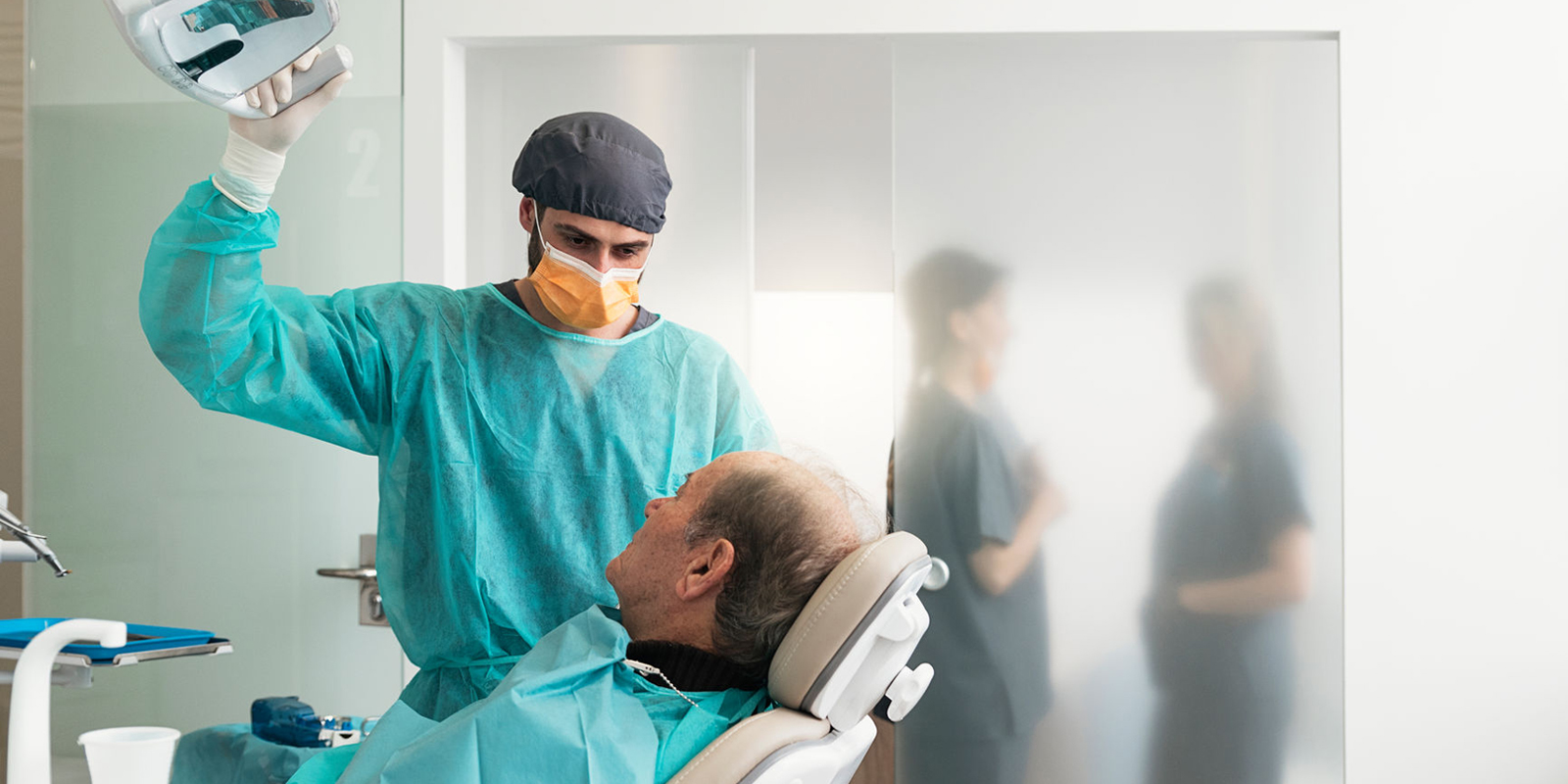 ACDQ dentist adjusting the lamp during an examination, with dental hygienists talking in the background